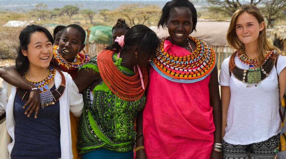 Young, female volunteers take a group photo with local Kenyan women. They are smiling, laughing and all wearing traditional Kenyan necklaces.