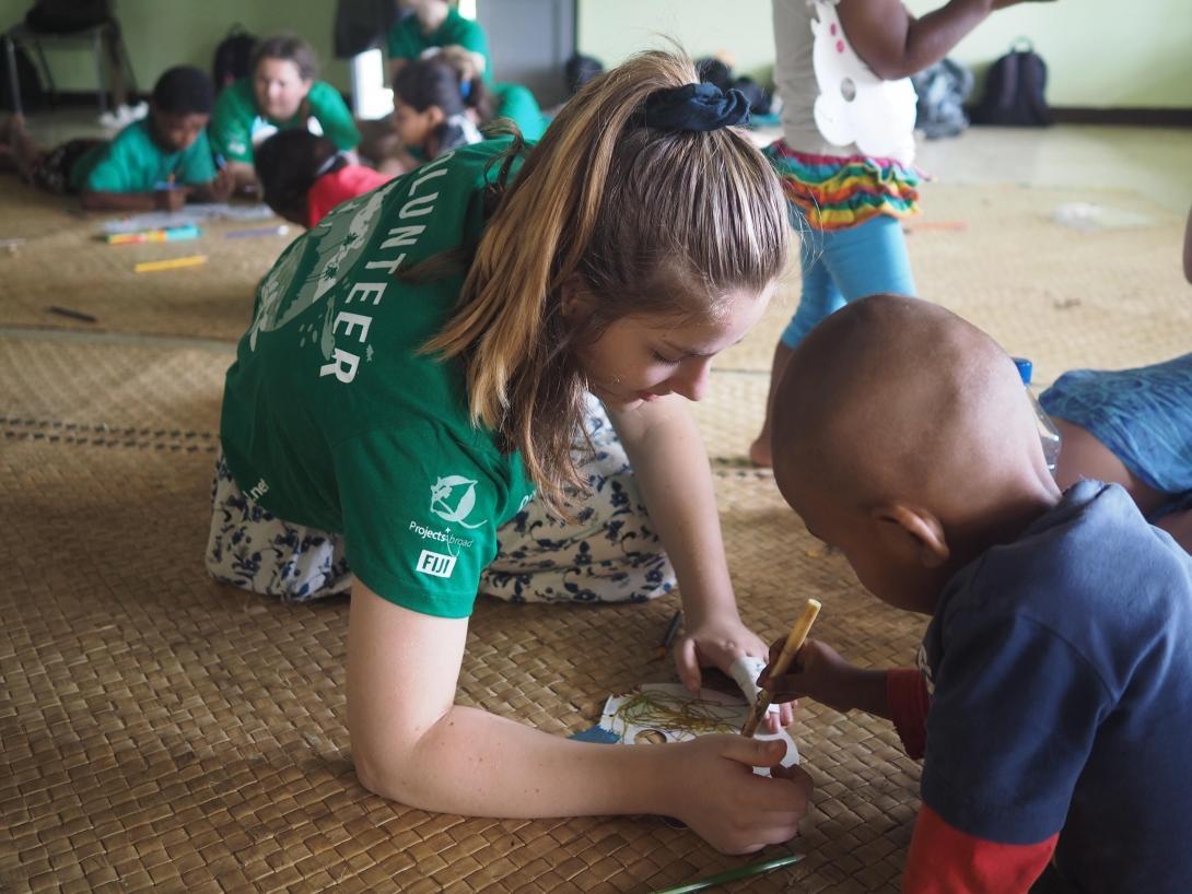 Projects Abroad volunteer with children in Fiji sits on the floor of a special needs day school to help a child to draw during her Childcare Project.