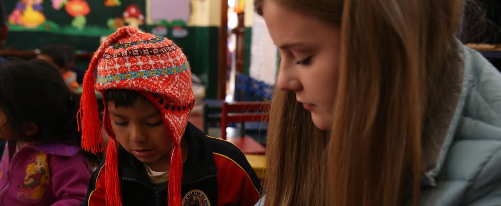 A volunteer working with children in Peru sits with a child in a classroom to help them with an activity.