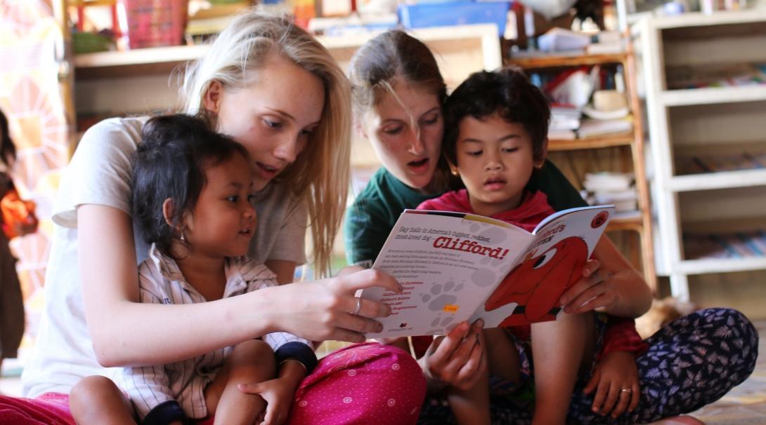 Children at a day care centre listen to a book being read by two female volunteers working with children in Cambodia.